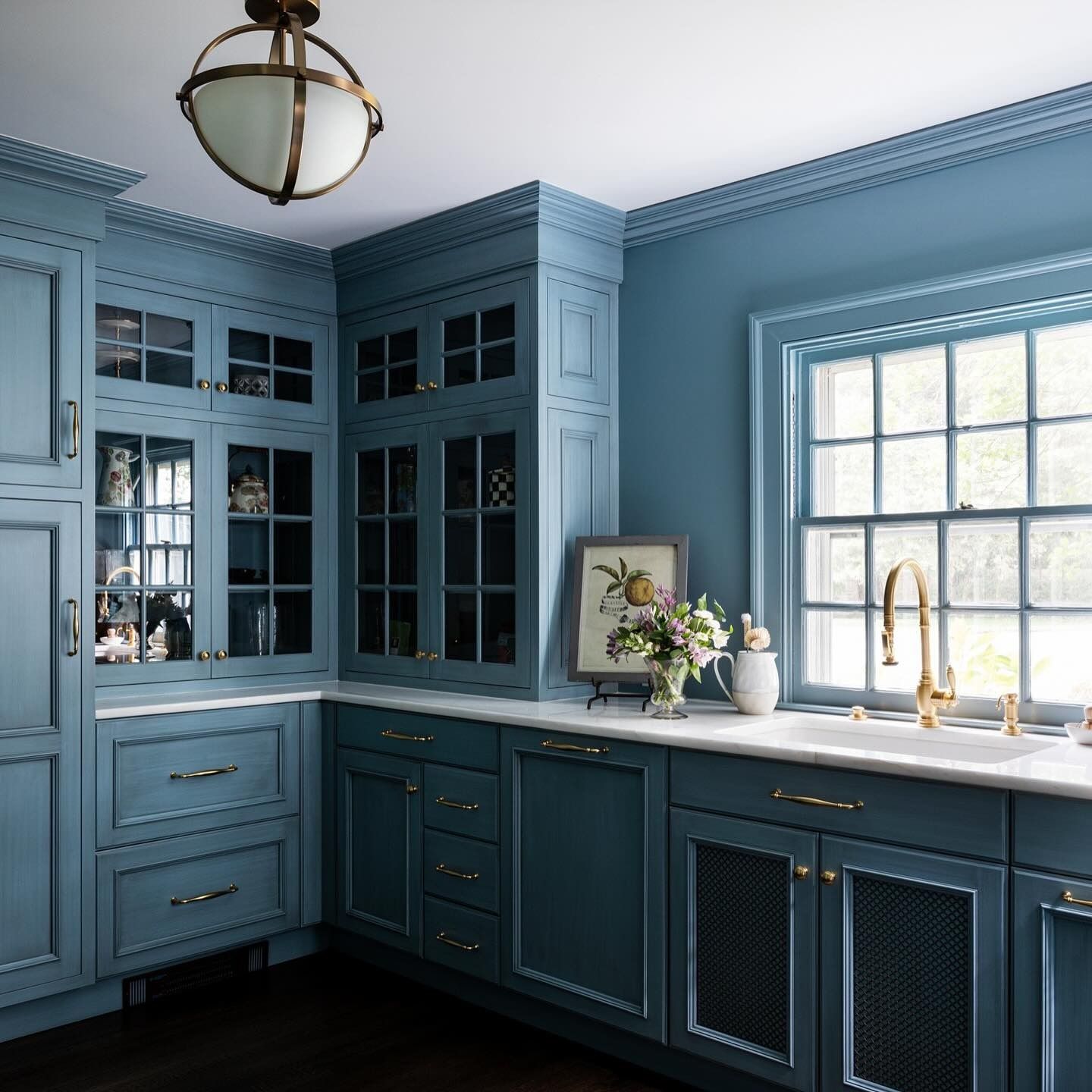 A kitchen with elegantly painted blue cabinets, brass hardware, and a bright window over the sink